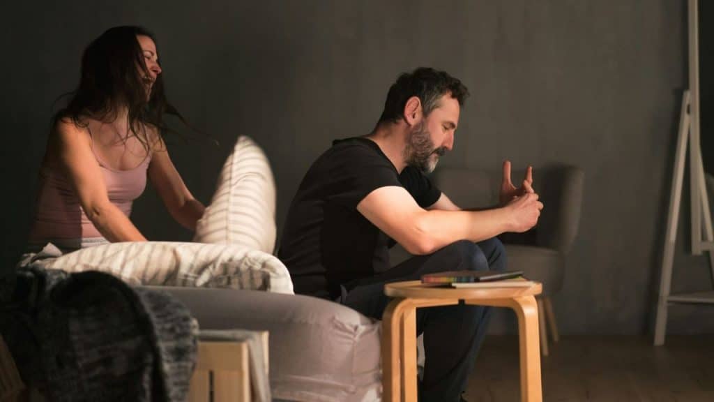 A frustrated man sits with his hands up while a yelling woman hits him with a pillow.