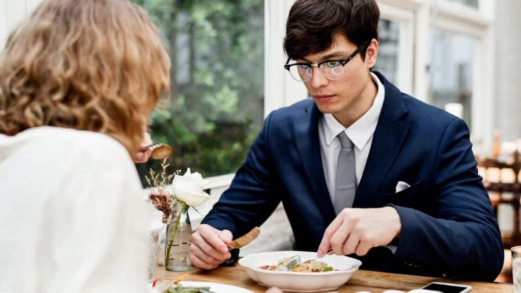 A man in a suit and glasses eats dinner across from a person in a restaurant.