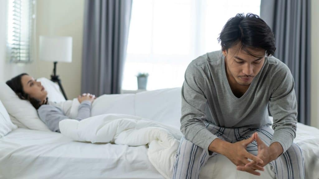A distressed man sits on the edge of a bed in a bright bedroom, while a woman lies asleep or unresponsive behind him.