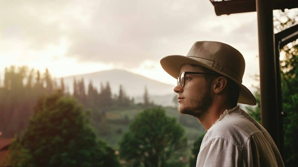 A side profile of a young man wearing a fedora and glasses, looking thoughtfully toward a view of misty forested mountains at sunset.