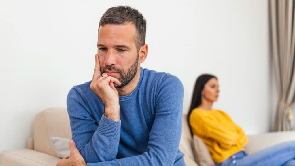 A focused image of a pensive man on a couch, with a woman sitting away from him in the background