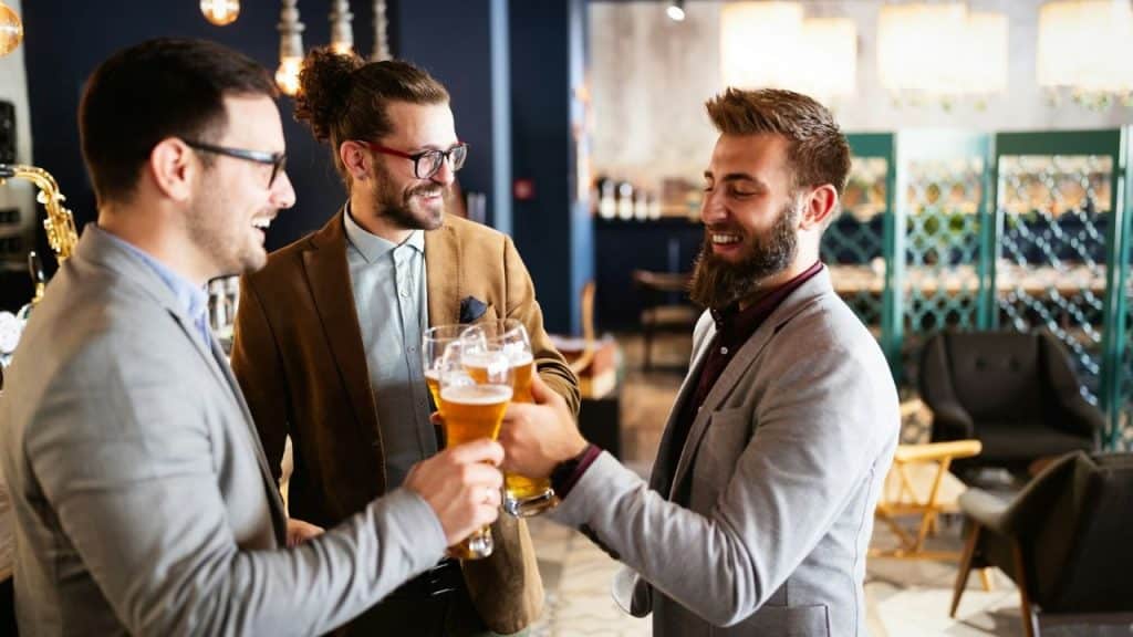 Three smiling men in business casual attire toast each other with beers in a bar.