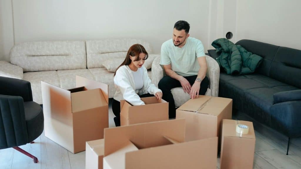 A smiling man watches a woman unpack cardboard boxes in a living room.