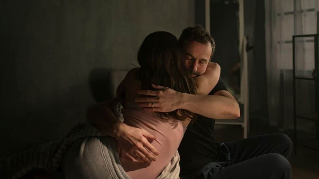 A man with his eyes closed sits on the bed, embracing a woman tightly in a dimly lit room.