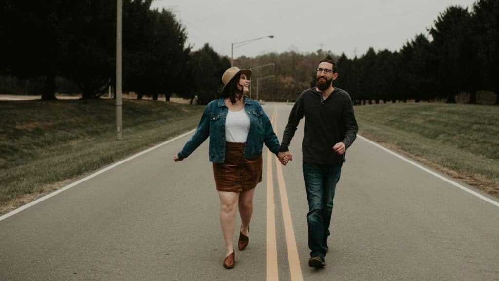 A smiling couple holding hands walks down the middle of an empty, asphalt road.