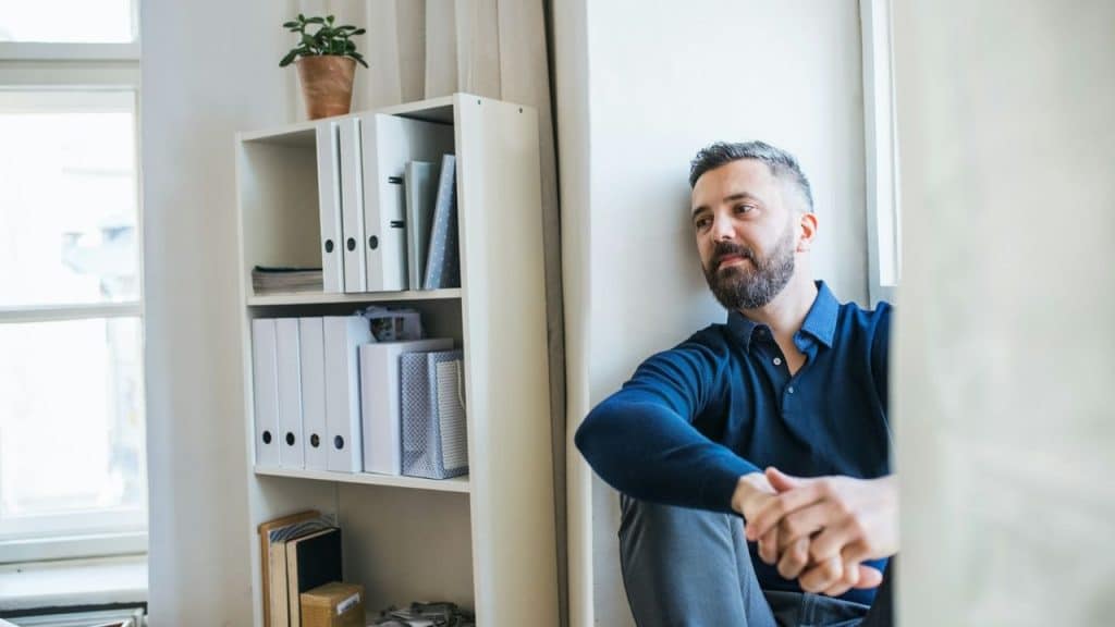 A bearded man in a blue shirt sits on the floor next to a white shelf and a window.