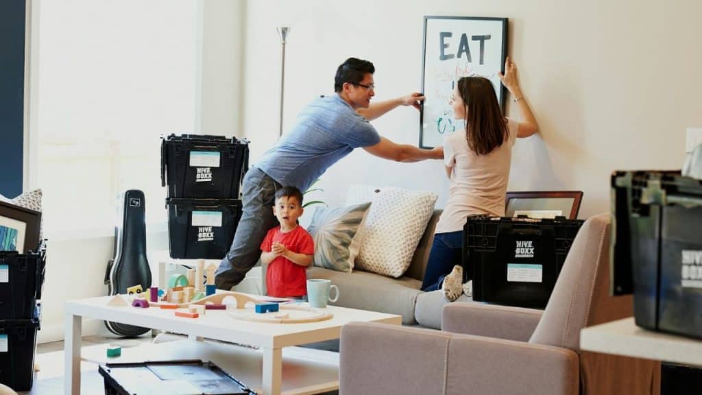 A couple hangs a picture while a young child watches them in a living room with moving boxes.