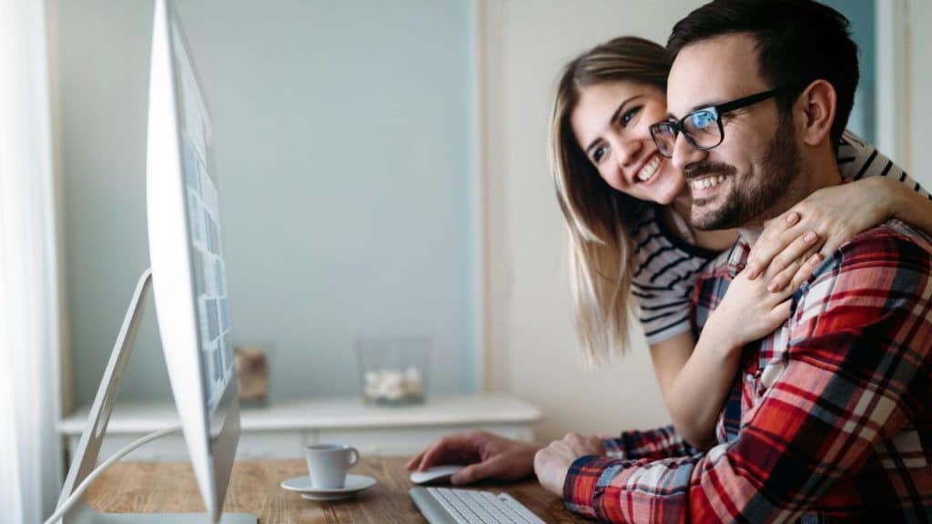 A man and a woman smile at a computer screen, with the woman embracing the man.