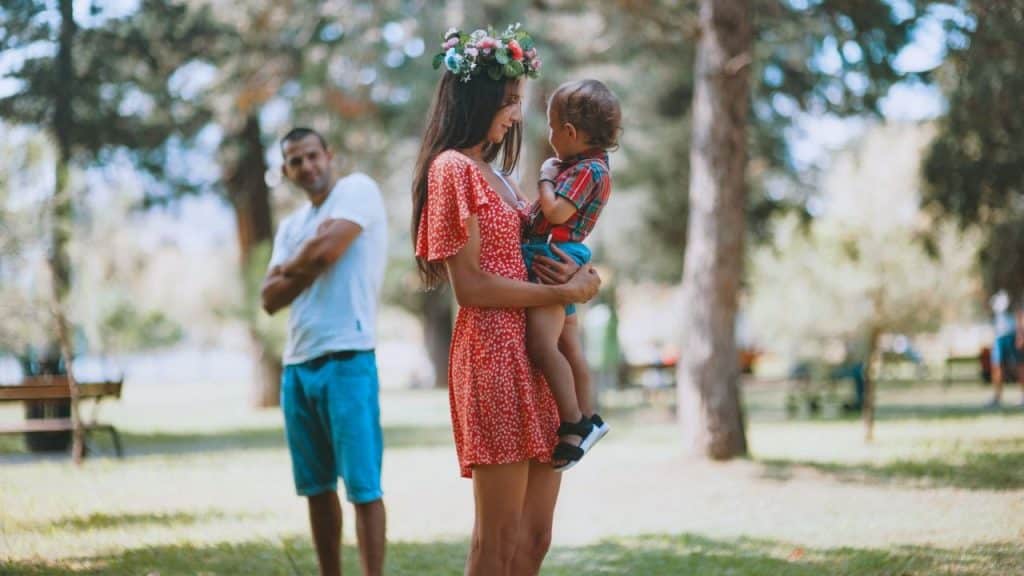 A woman holding a child looks at him; a man with crossed arms is blurred in the background.