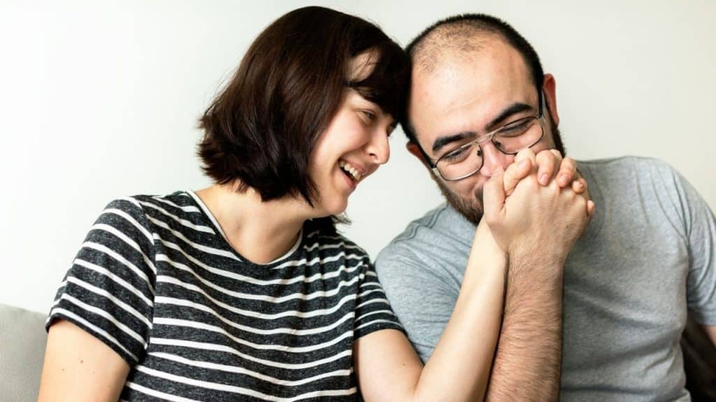 A couple smiles and holds hands, pressing their heads close together against a white wall.