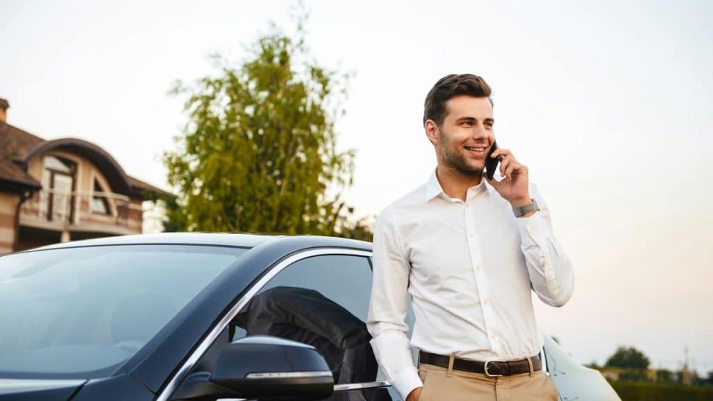 A man in a white shirt talking on the phone next to a parked car