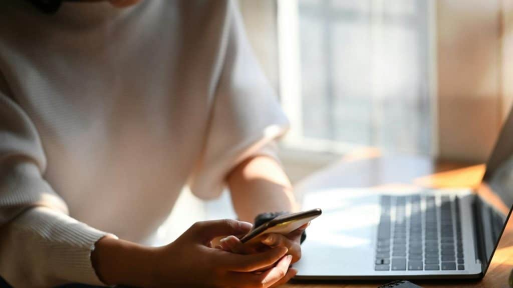 A person sitting at a desk using a smartphone beside an open laptop.