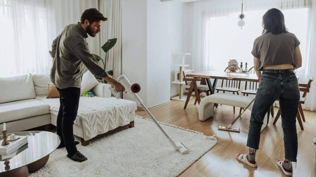 A man and woman vacuum a rug in a bright, modern living room and dining area.