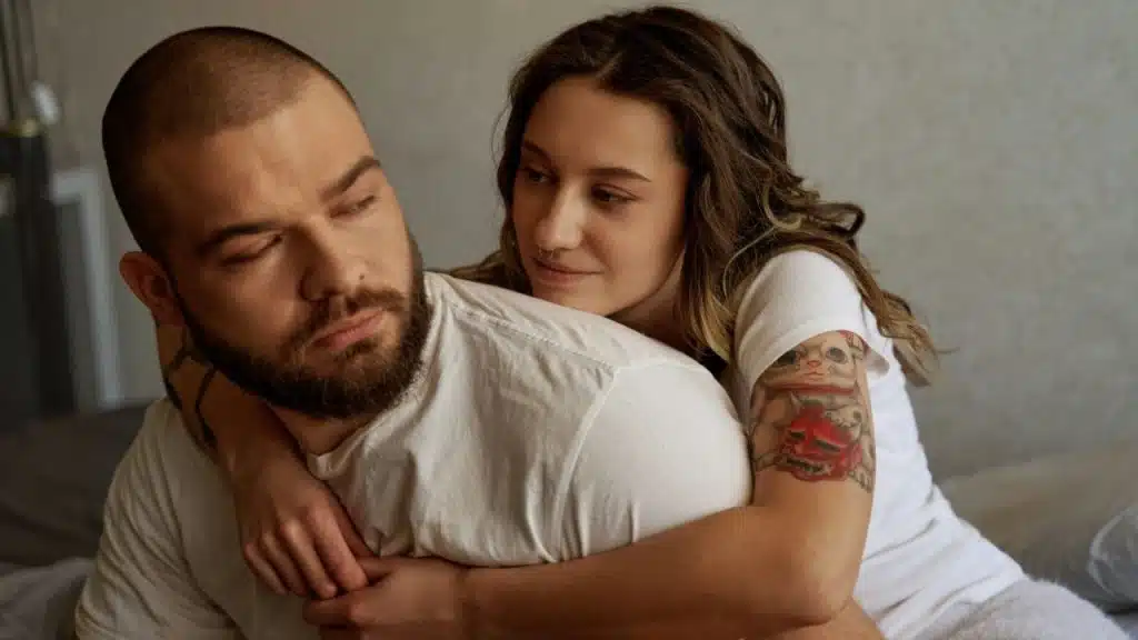 Woman with tattoos and a nose piercing hugging a bearded man from behind.