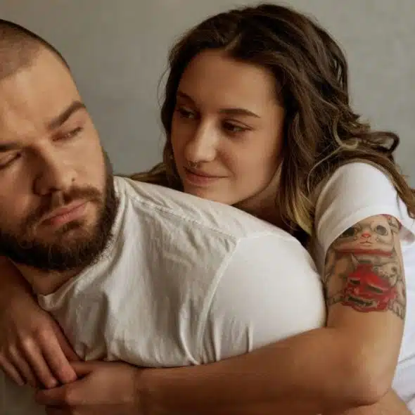 Woman with tattoos and a nose piercing hugging a bearded man from behind.