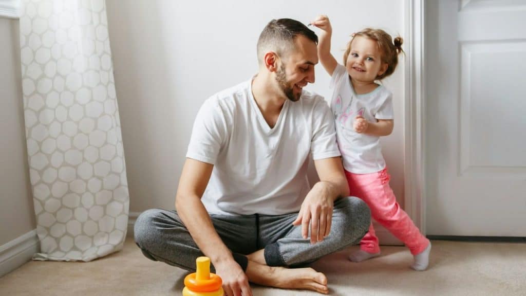 Father in a white shirt sitting on the floor with a toddler smiling and playing.