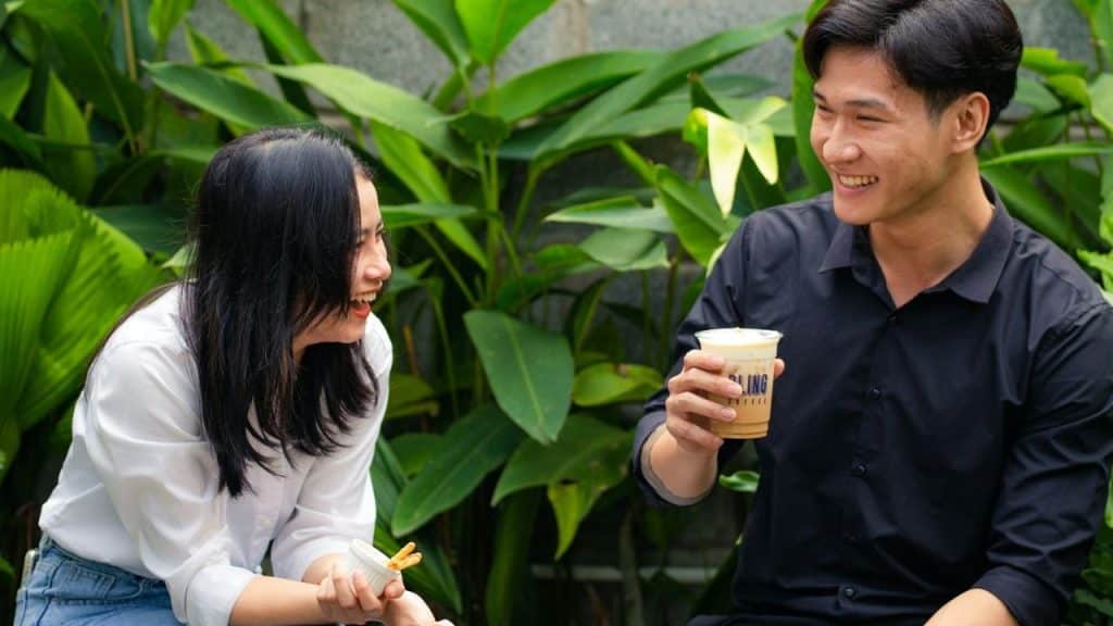 Two smiling people outdoors with drinks, surrounded by large green leaves.