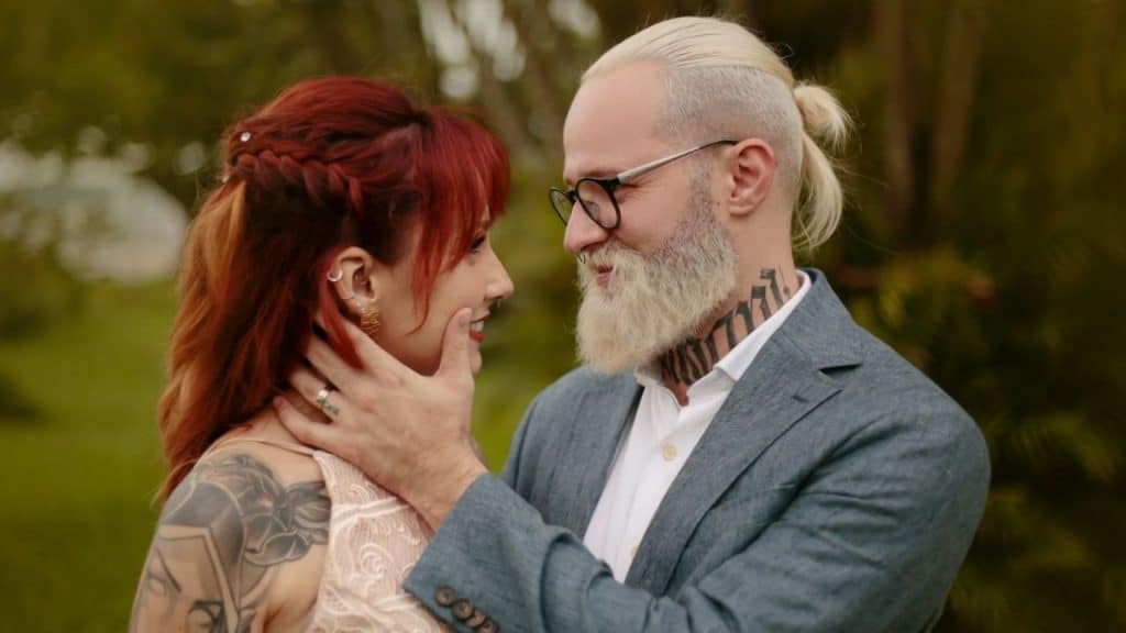 Man with a blonde beard and ponytail gently touching a red-haired woman's face.