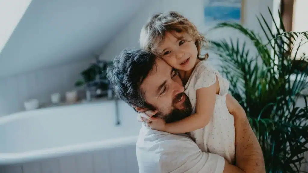 Father with a beard hugging a young child in a light-filled room with a bathtub.