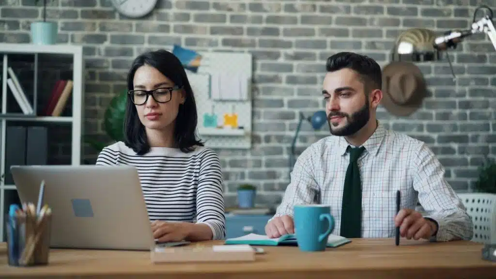 Two people working side-by-side at a desk with a laptop and a blue coffee mug.