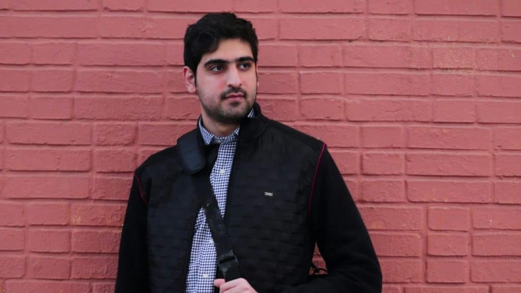 Young man in a black vest and patterned shirt standing against a red brick wall.