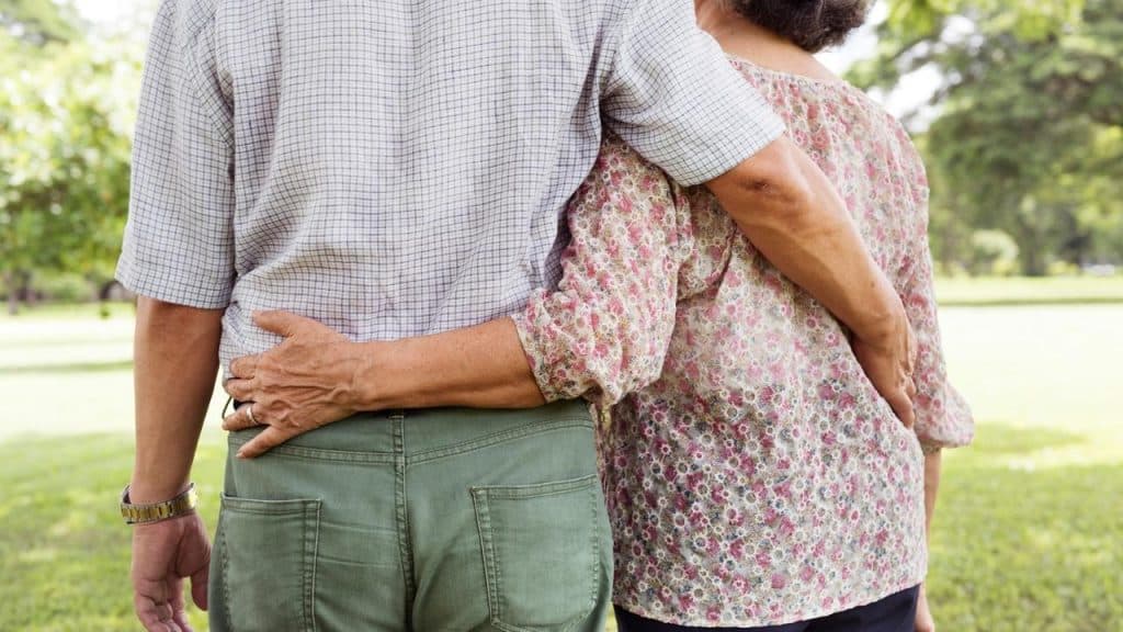 An older couple stands arm in arm together in a sunny park.