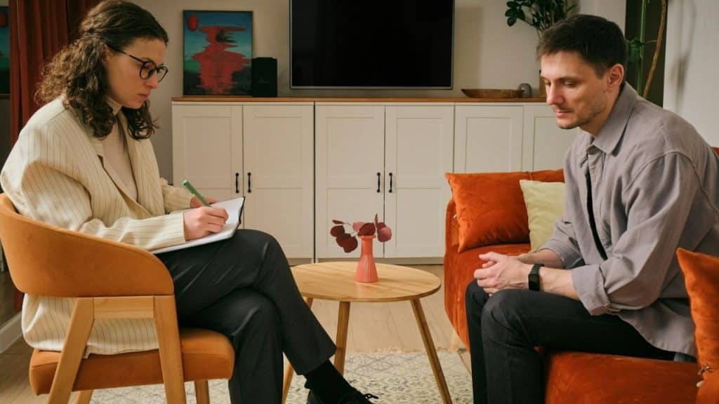Woman taking notes across from a man sitting on an orange couch in a living room.