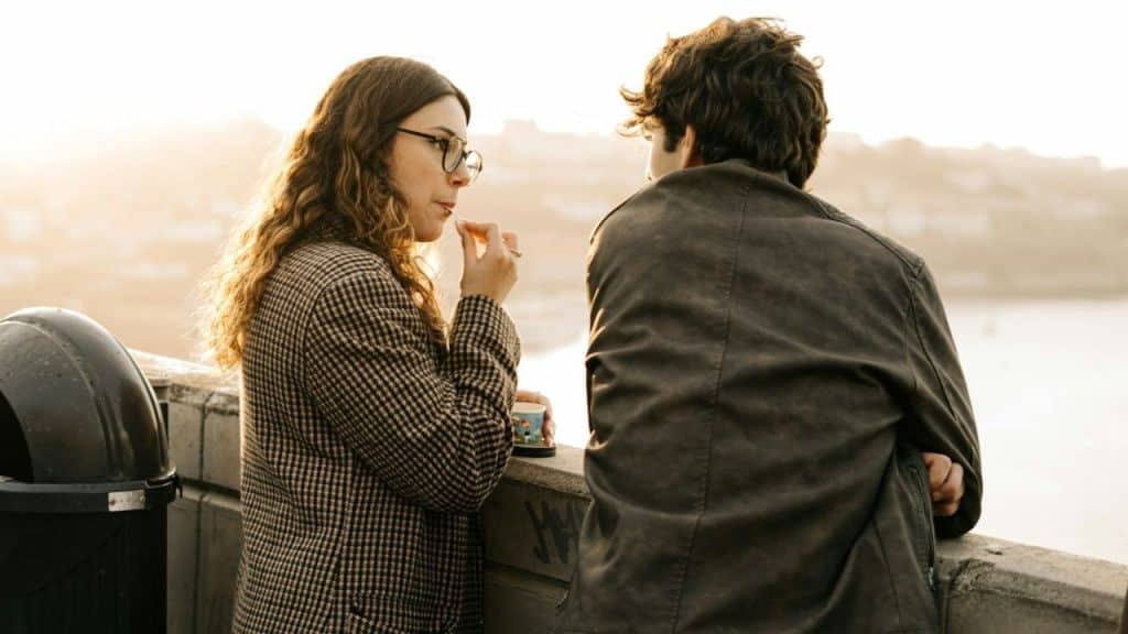 Two people talking outside at sunset, leaning on a wall, with a woman holding a mug.