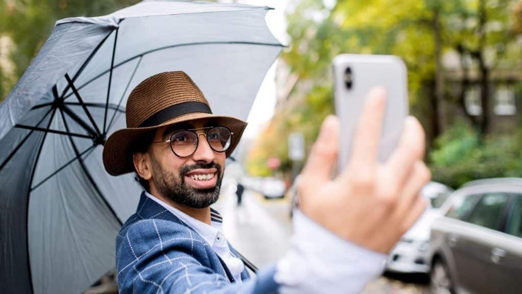 Smiling man in a fedora, glasses, and blazer taking a selfie under an umbrella outdoors.