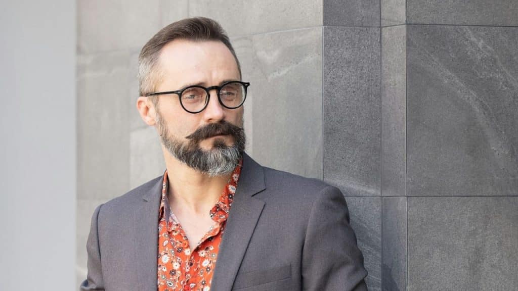 Man in a gray blazer and orange patterned shirt with a beard and glasses.