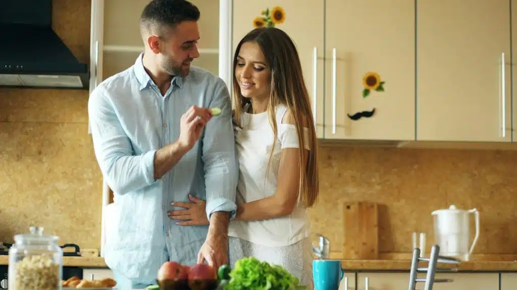 A man feeding his wife while working in the kitchen.