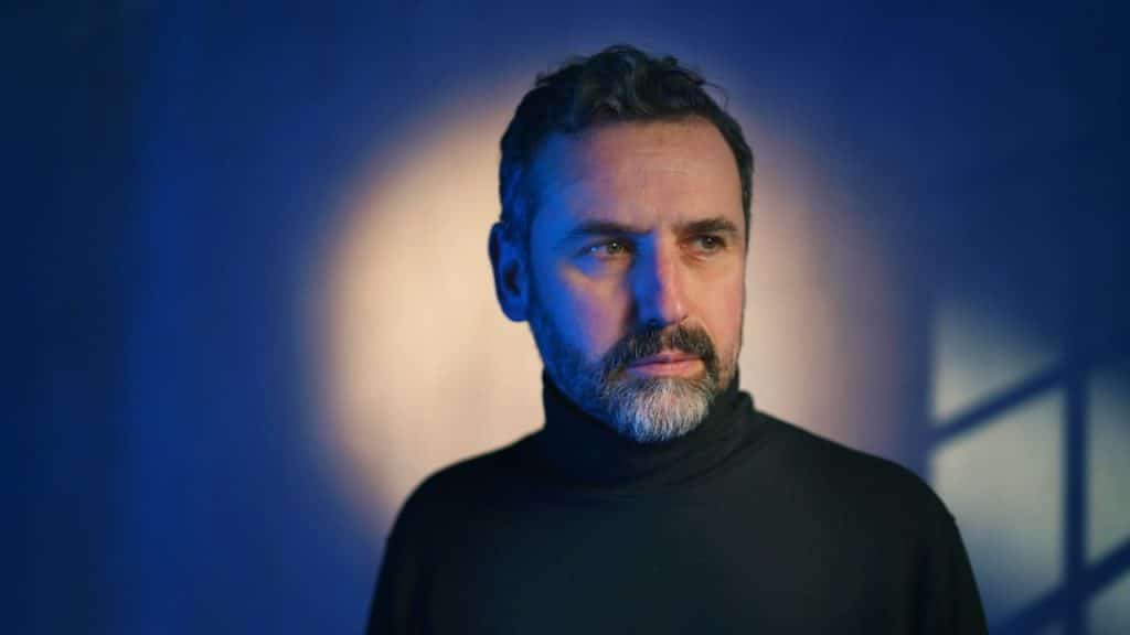 Man in a black turtleneck against a dark blue background with a white spotlight.