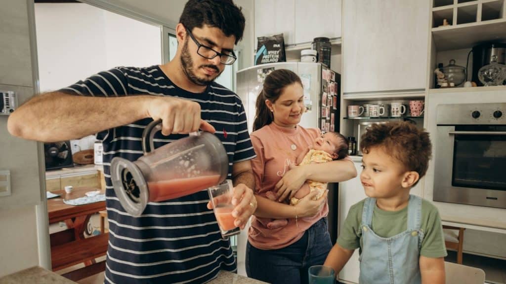 A man pours a drink for his son in a kitchen while his wife is holding a baby.