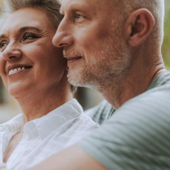 Close-up of a smiling older couple embracing and looking into the distance outdoors.