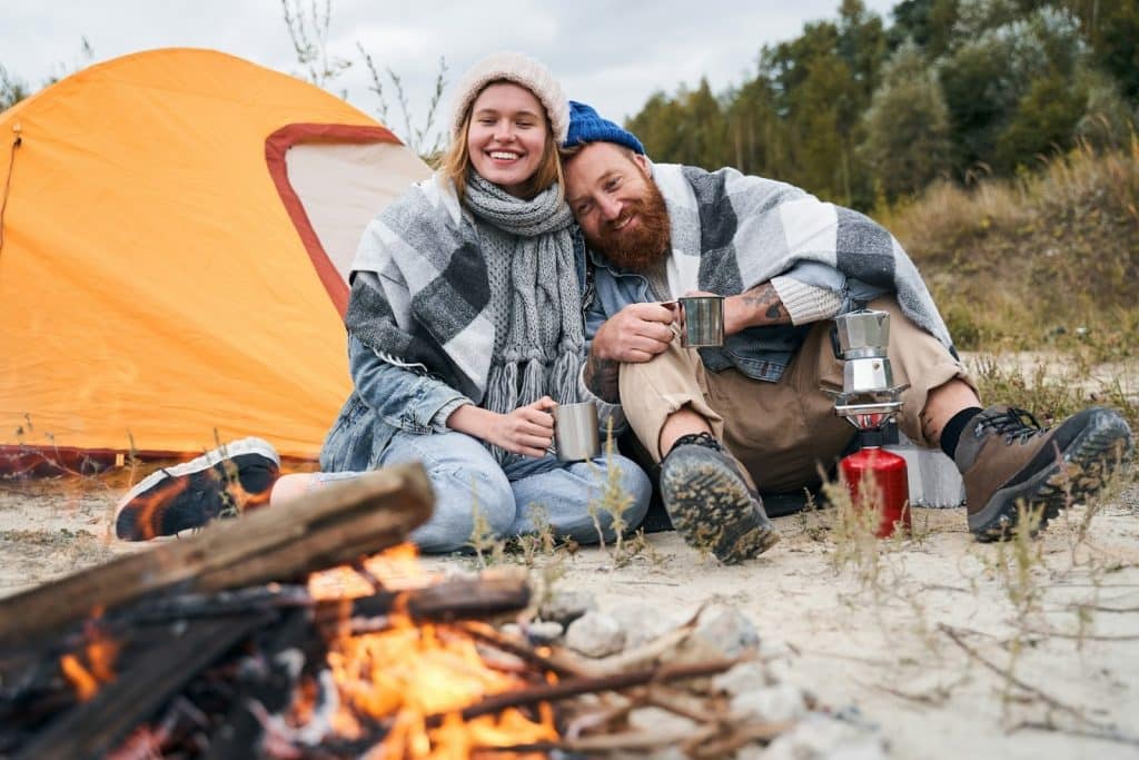 The couple is holding a cup and looking at the campfire.