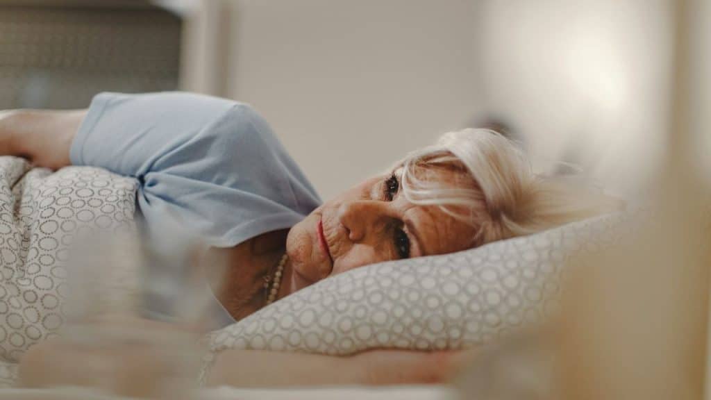 Older woman with white hair lying awake in bed and looking toward the camera.