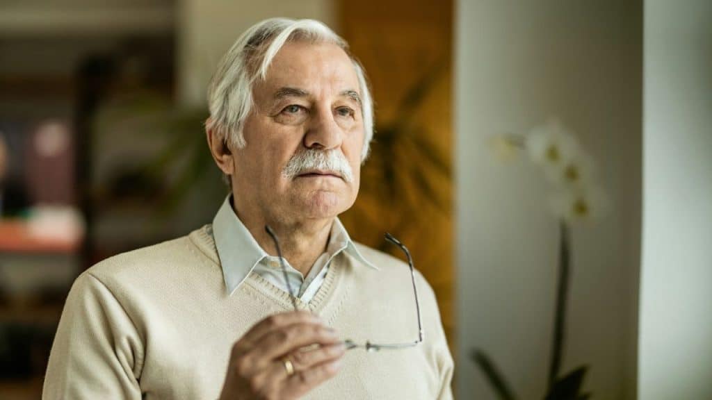 Older man with a mustache holding glasses, looking away thoughtfully indoors.