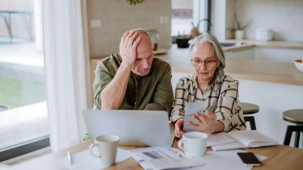 Older couple looking stressed while reviewing papers and a laptop at a kitchen table.