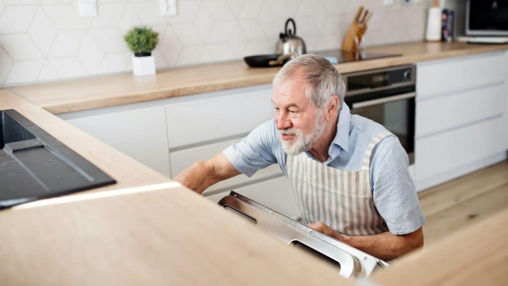 Older man in an apron working on a dishwasher or oven in a modern kitchen.