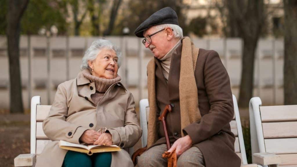 Older couple in coats and scarves sitting on a park bench, talking and smiling.