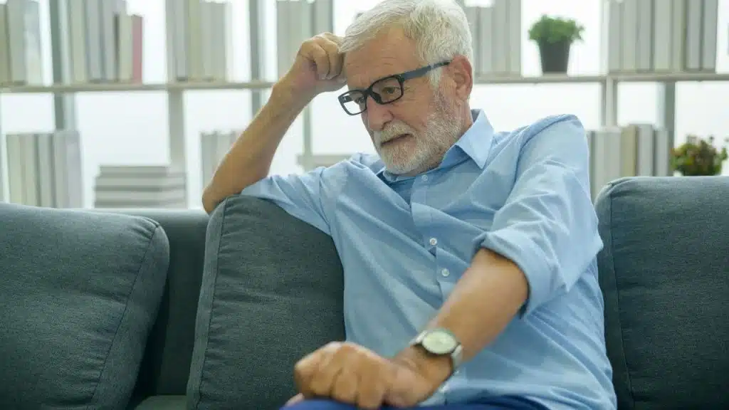 Older man with a beard and glasses sitting on a couch with a thoughtful expression.