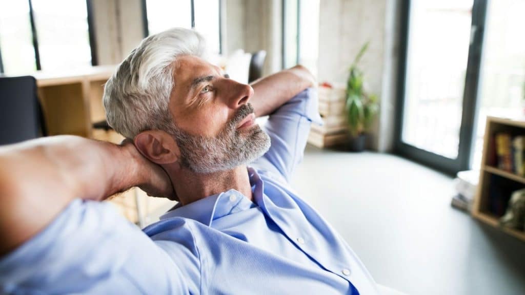 Middle-aged man with gray hair and beard relaxing in a chair with hands behind his head.