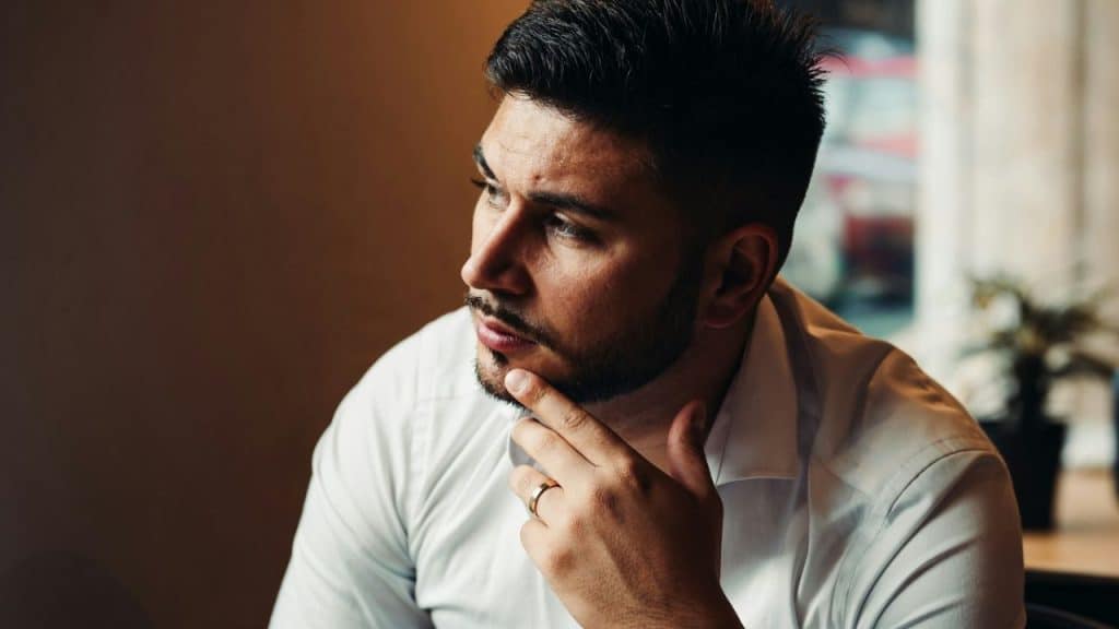 A serious man with a beard and a wedding ring sits contemplating with his hand on his chin.
