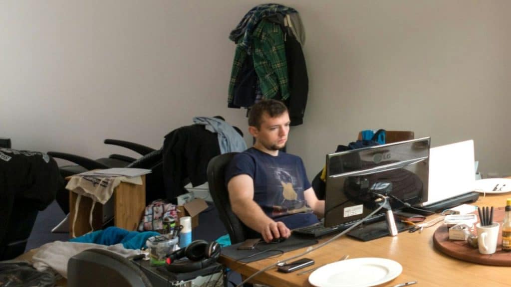A young man is sitting at a computer in a messy room with clothes piled up and a monitor on a wooden desk.
