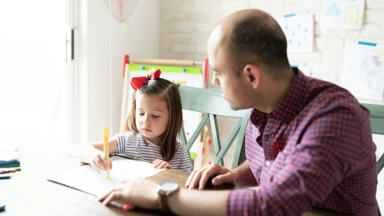 Father and daughter sit at a table coloring with markers on a white paper.