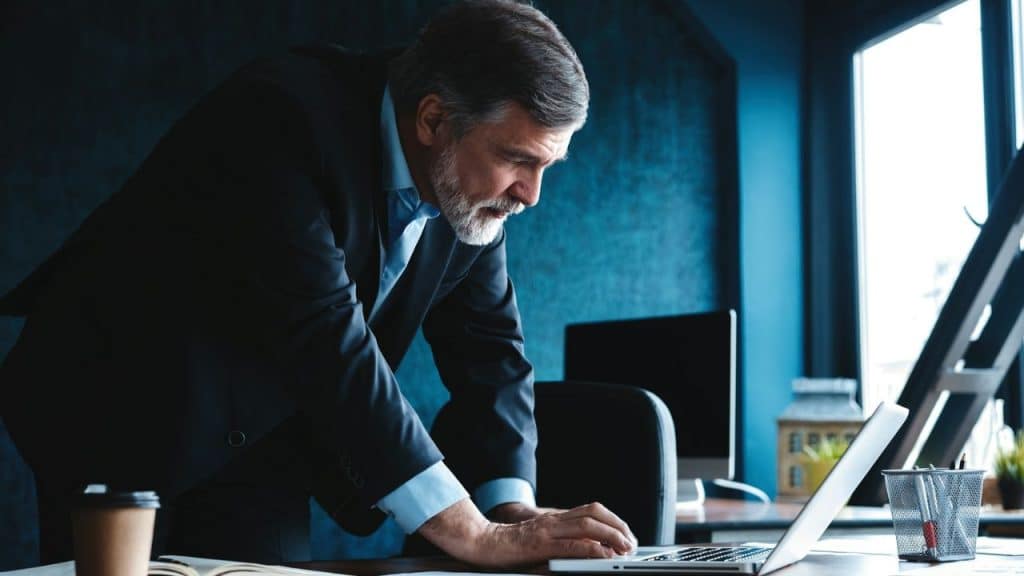 Senior man in a dark suit leaning over his desk, working on a laptop.