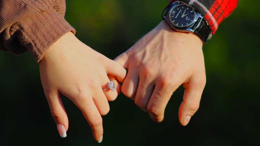 A close-up of a man's and a woman's hands intertwined by their pinkies, with the woman wearing an engagement ring.