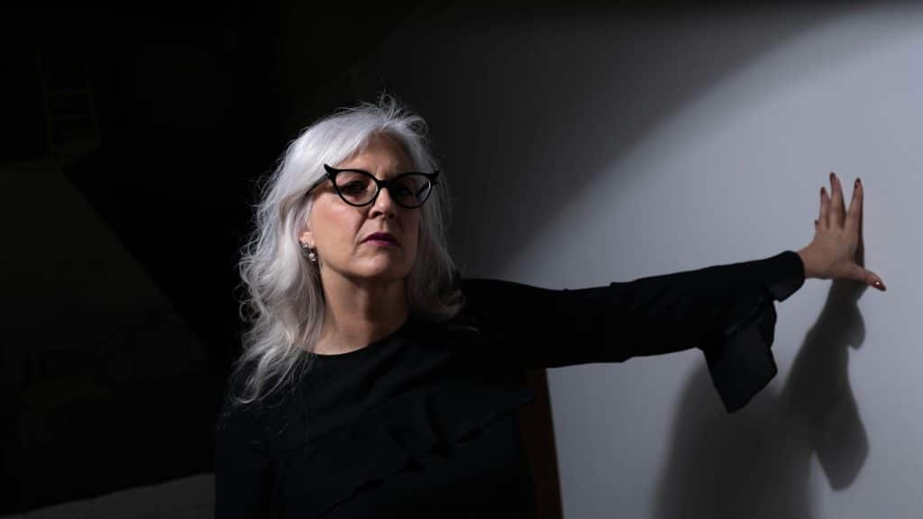An older woman wearing glasses is leaning on a wall.
