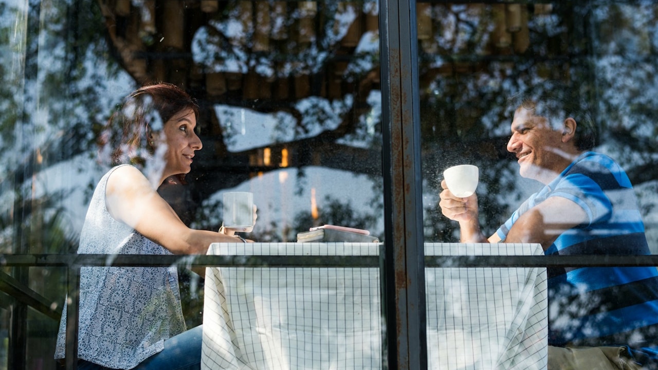 A smiling man and woman sit opposite each other at a cafe table, holding coffee cups, viewed through a glass window.