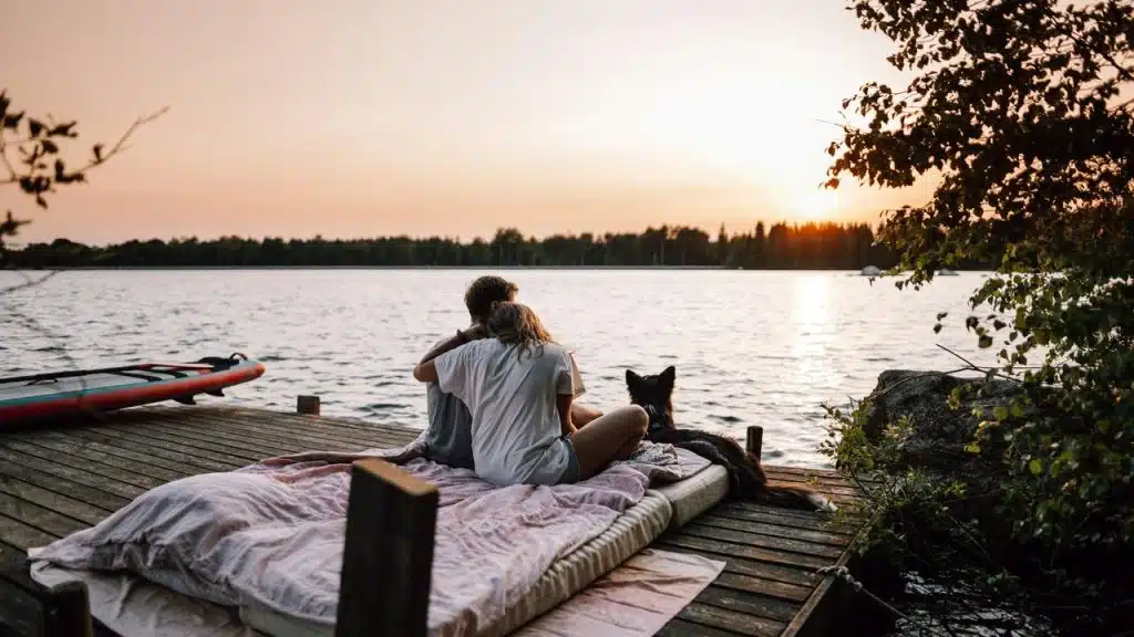 A couple sitting with their dog on a dock watching the sunset over a lake.
